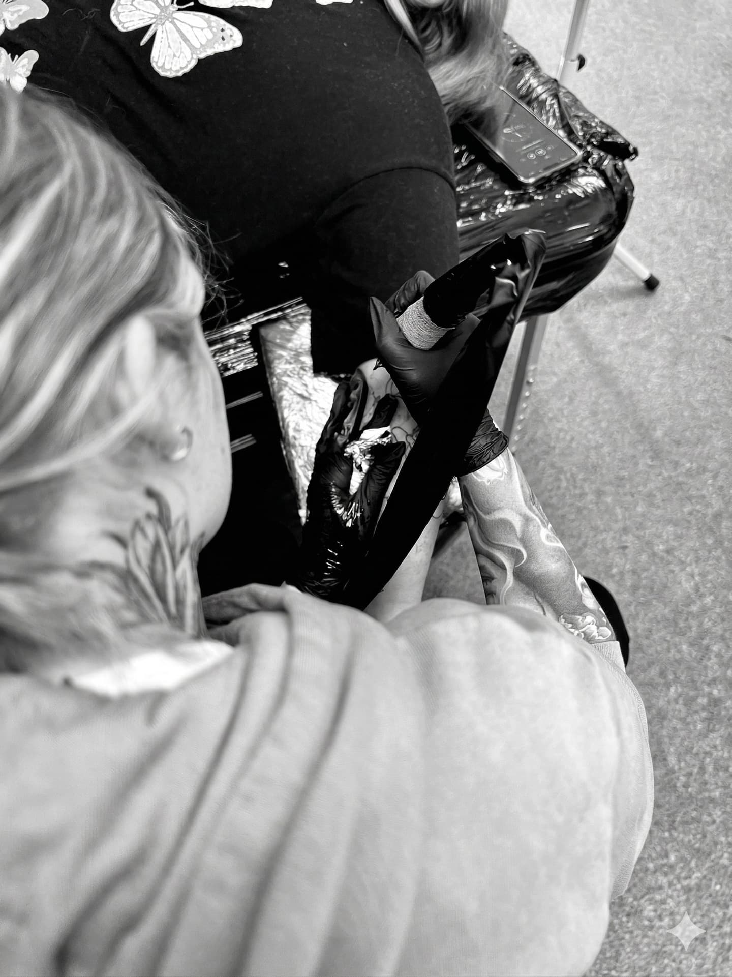 A black and white close-up photo of a tattoo artist from Emmy Tattoo working on a client's arm using a rotary tattoo machine.
