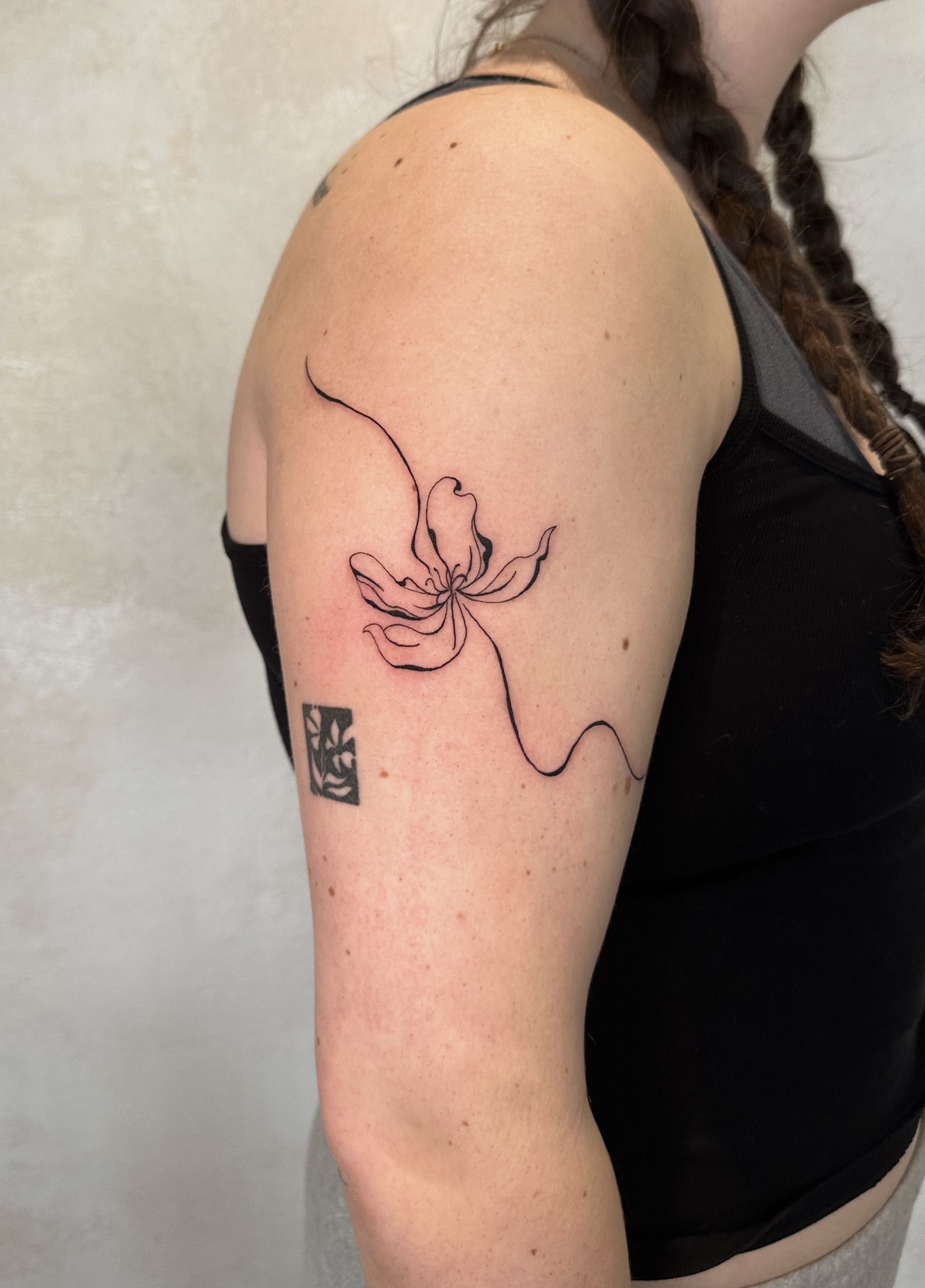 Minimalist fine-line tattoo on an upper arm featuring an abstract, continuous flowing line that forms a delicate flower.