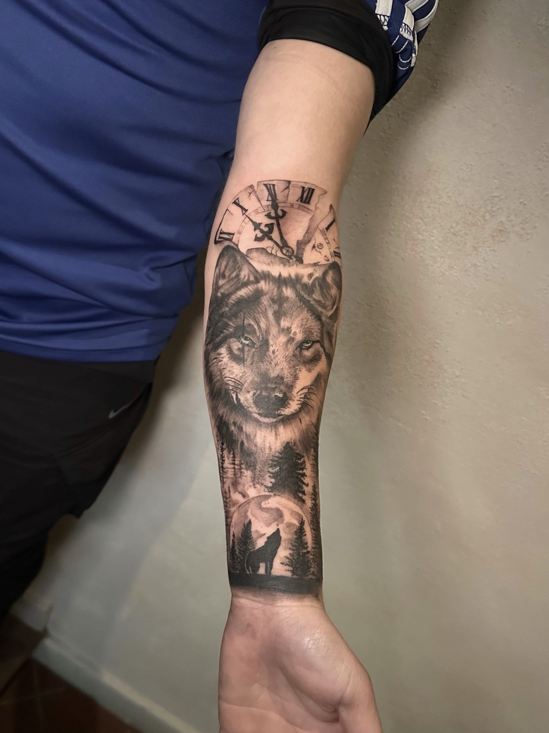 Black and grey forearm half-sleeve tattoo featuring a realistic wolf face, a shattered Roman numeral clock, and a forest silhouette with a howling wolf.