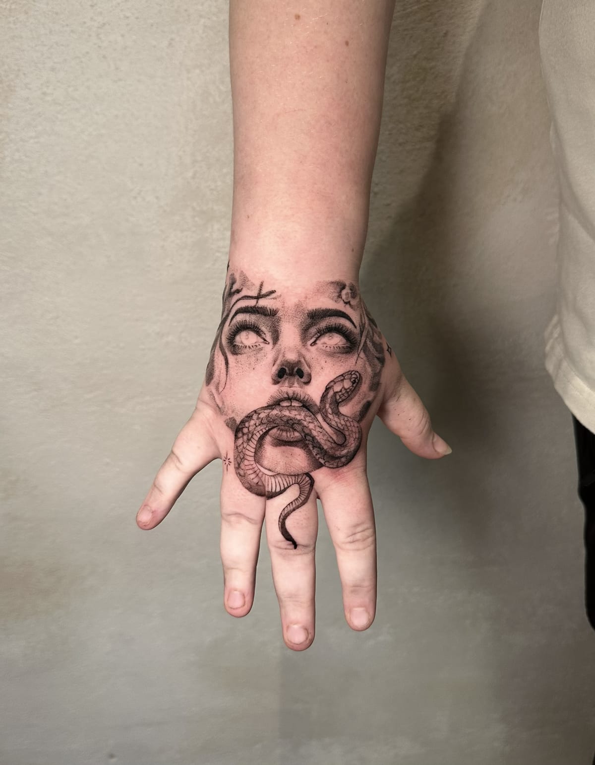 Black and grey surrealism hand tattoo featuring a woman's face with blank eyes and a snake winding through her mouth, designed by Emmy Tattoo.