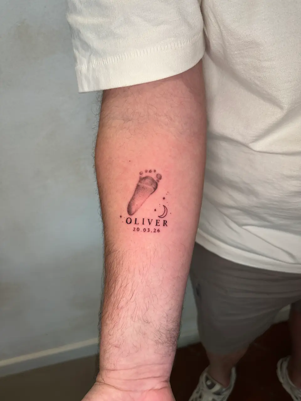 Shaded baby footprint above a name and date, with a small crescent moon on the forearm.