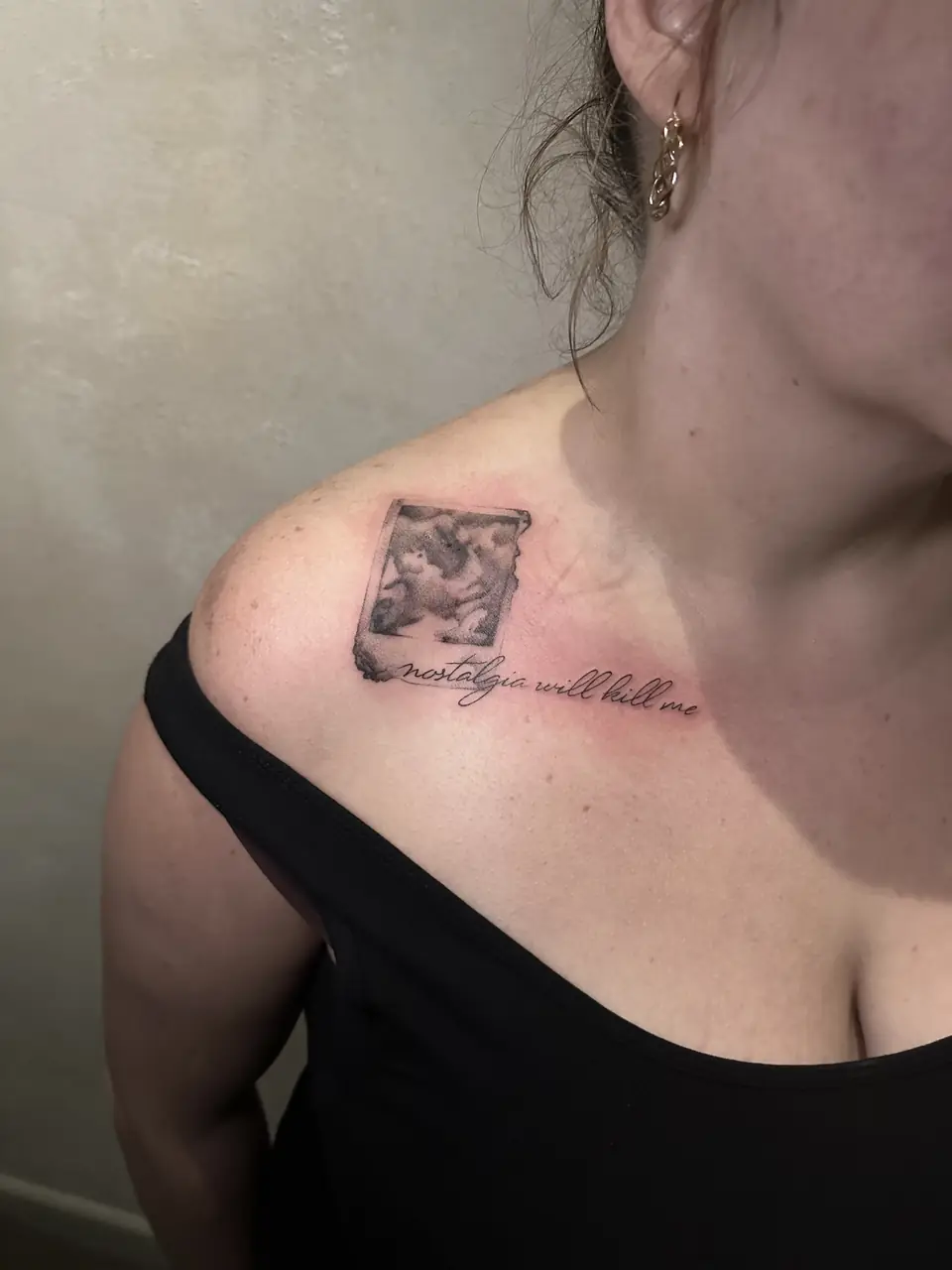 Small black and grey cherub painting with cursive lettering beneath it near the collarbone.