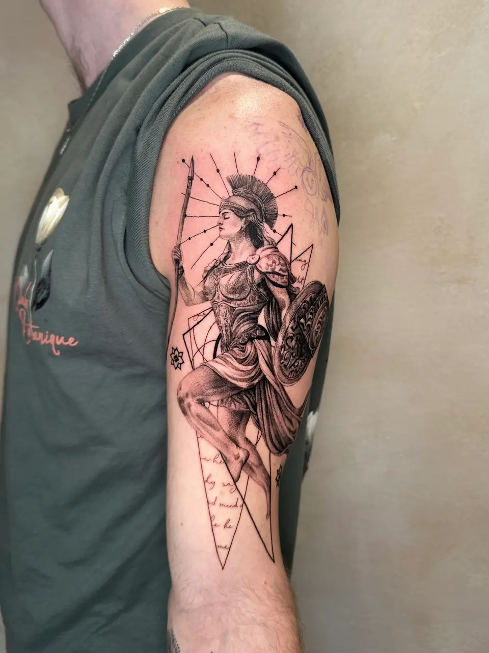 Classical helmeted warrior with a spear and decorated shield over geometric linework on the upper arm.