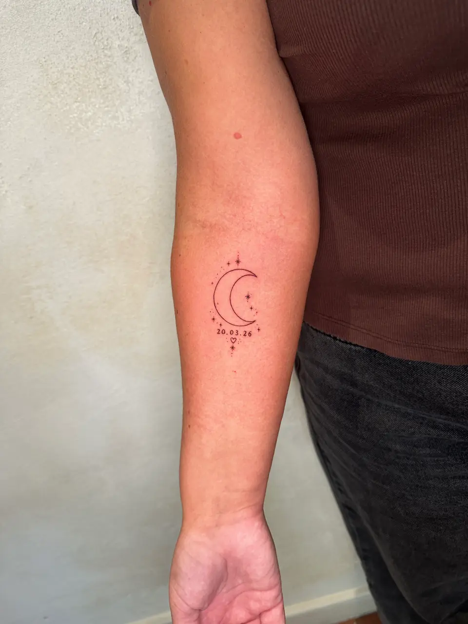 Fine-line crescent moon with small stars, dots and a date on the forearm.