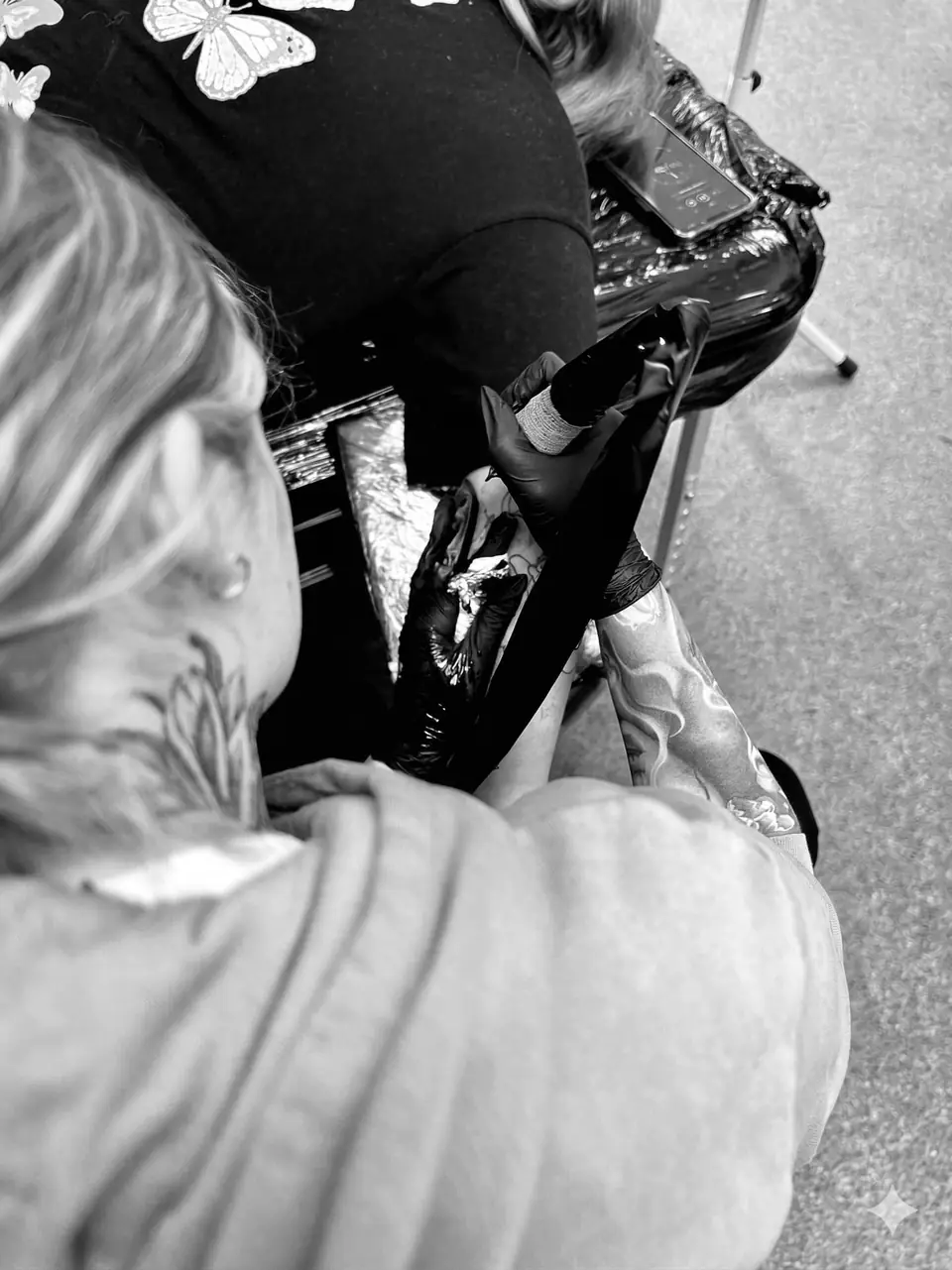 A black and white close-up photo of a tattoo artist from Emmy Tattoo working on a client's arm using a rotary tattoo machine.