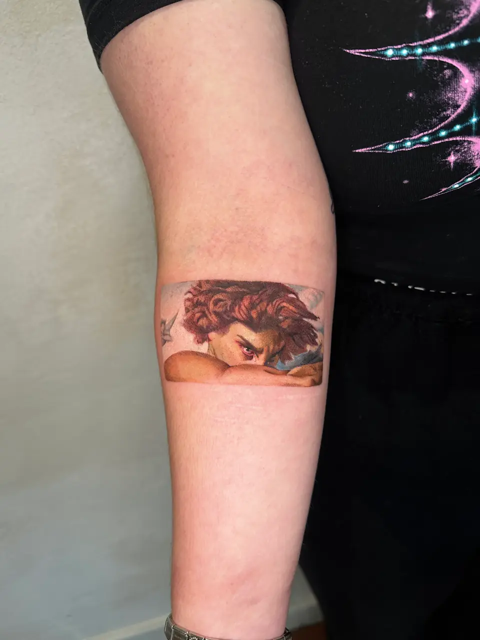 Colour painting detail of the Fallen Angel's eyes above a bent arm, tattooed in a rectangular panel.