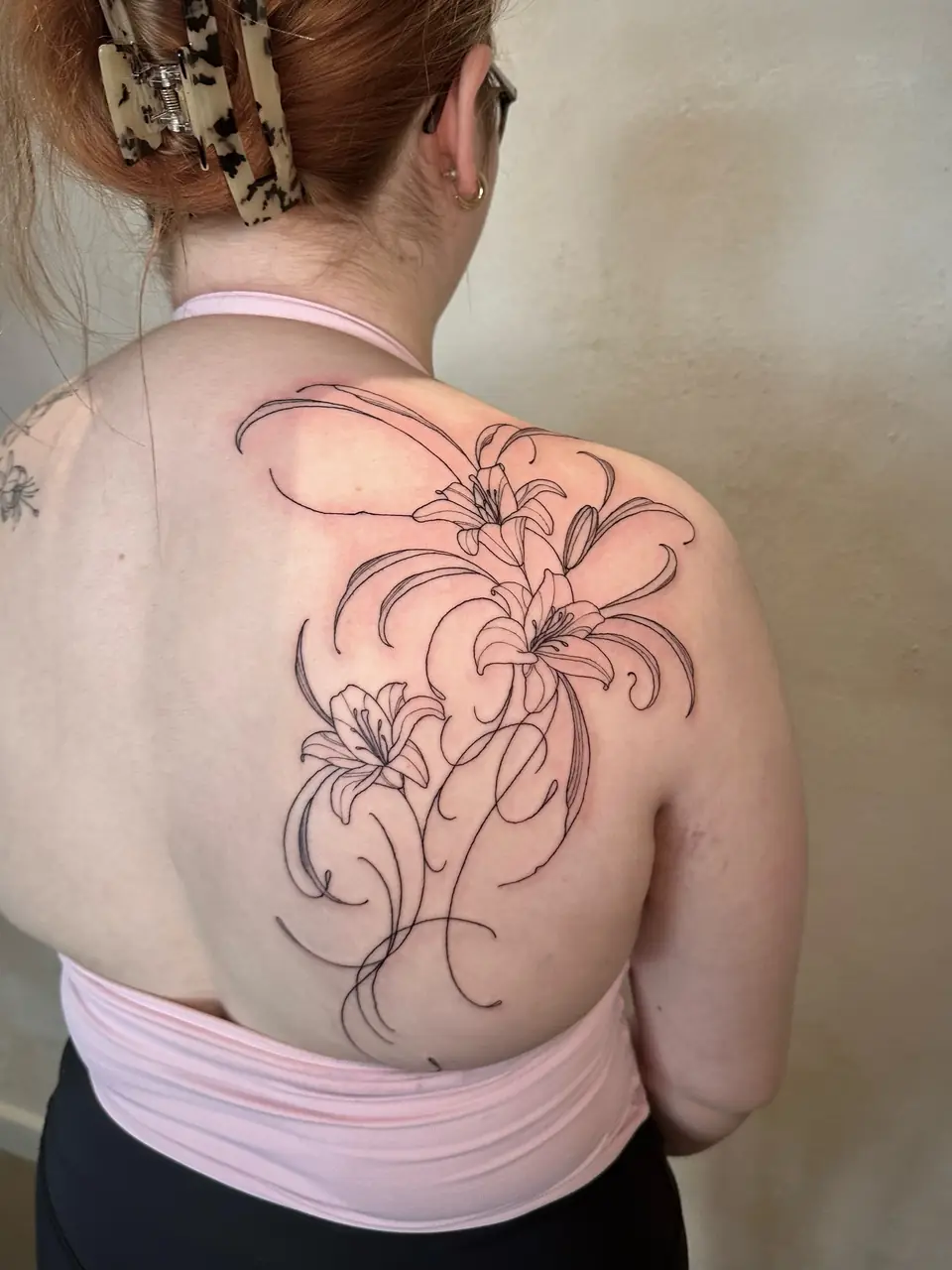Three fine-line lilies with long curling petals and stems across the shoulder blade.