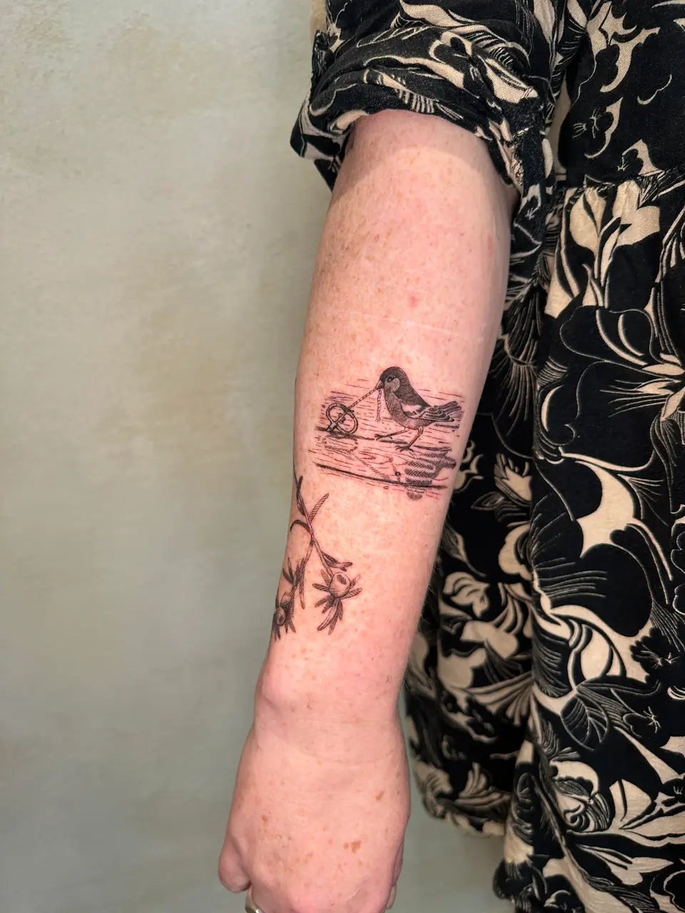 Black etched-style bird holding a ring on a cord in a small scene on the forearm.