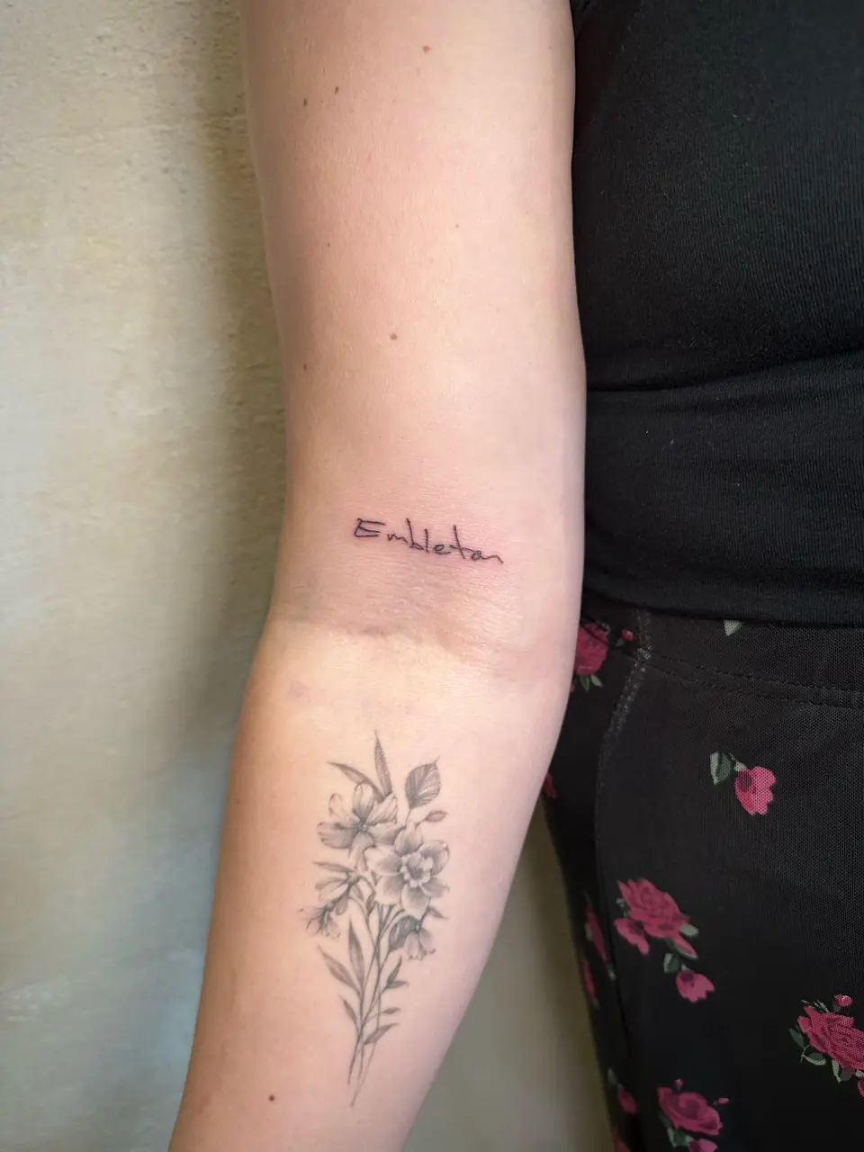 Small handwritten word above the inner elbow, with a floral tattoo visible below.