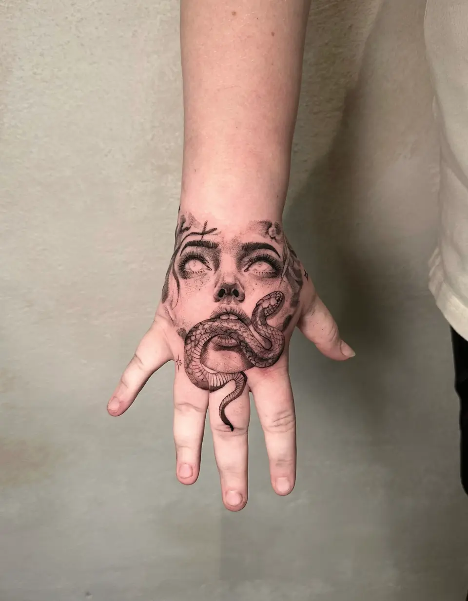 Black and grey woman's face with blank eyes and a snake winding across the mouth on the back of the hand.